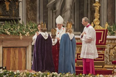 Drei Sternsinger stehen vor Papst Franziskus. Sie haben die traditionellen Gewänder an. Die Sternsinger haben an der Gabenprozession teilgenommen. Der Papst blick sie gütig und dankbar an. Er steht direkt vor ihnen und sitzt nicht auf seinem Goldenen mit rotem Samt bezogenen Stuhl.