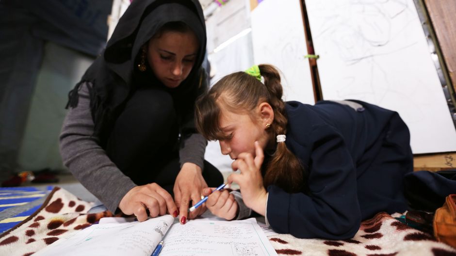 Khadija und ihre Mutter sitzen auf dem Boden und gucken in ein Arbeitsheft von Khadija. Ihre Mutter hilft Khadija bei den Schulaufgaben.