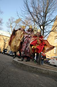 Eine Sternsingergruppe springt von einer Stufe herunter und strahlt viel positive Energie aus. Die traditionellen Gewänder bewegen sich dynamisch im Schwung. Sie tragen goldene Kronen und haben auch in der Luft den Stern der Sternsinger fest im Griff.