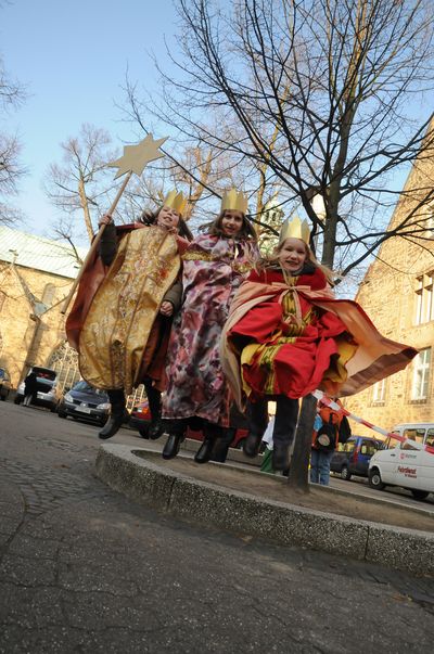 Eine Sternsingergruppe springt von einer Stufe herunter und strahlt viel positive Energie aus. Die traditionellen Gewänder bewegen sich dynamisch im Schwung. Sie tragen goldene Kronen und haben auch in der Luft den Stern der Sternsinger fest im Griff.