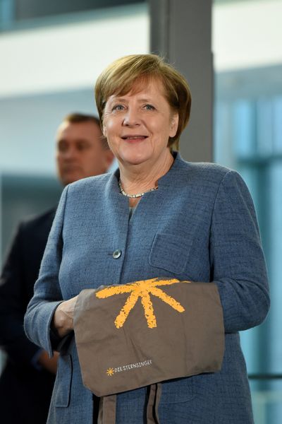 Die Bundeskanzlerin Angela Merkel mit einem Jutebeutel der Sternsinger. Den Beutel hat sie sich über den linken Unterarm gelegt und den Arm leicht angewinkelt. 