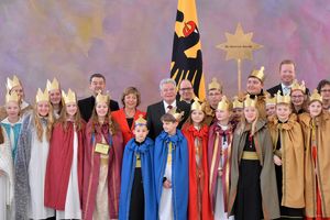 Neunzehn Sternsinger stehen in einer Gruppe zusammen mit Bundespräsident Joachim Gauck, seiner Lebensgefährtin und Prälat Krämer. Im Hintergrund ist die Nationalflagge von Deutschland zu sehen. Die Sternsinger tragen Umhänge in Rot, Blau und Grün. Alle Sternsinger tragen goldene Kronen. Im Hintergrund hält einer der Sternsinger den Stern von Betlehem in die Luft.