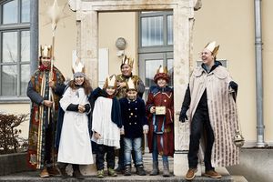 Abschlussfoto der Sternsingergruppe: Sieben Sternsinger stehen vor einem Gebäude und lachen. Ein Mädchen trägt einen Stern, ein Junge schwenkt ein Weihrauchfass und ein anderes Mädchen hat die Sammeldose in der Hand.