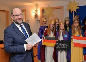 Eu- Parlamentspräsident Martin Schulz freut sich über den Besuch der Sternsinger. Er hält ein Notenheftchen in der Hand und lächelt in die Kamera. Im Hintergrund stehen die Sternsinger und singen ein Lied. Die Sternsinger haben festliche Kleidung an und tragen Kronen auf dem Kopf.