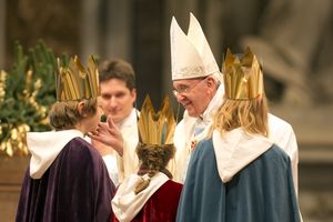 Drei Sternsinger stehen vor Papst Franziskus. Sie haben die traditionellen Gewänder an. Die Sternsinger haben an der Gabenprozession teilgenommen. Der Papst steht direkt vor den Sternsinger und blickt Ihnen und ie Augen. Er berührt einen der Sternsinger sanft mit der Hand an der Wange. 