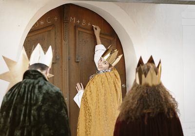 Drei Sternsinger schreiben den traditionellen Segen der Sternsinger an eine Haustür. Die Sternsinger tragen Kronen, den Stern und die traditionellen Gewänder der Heiligen Drei Könige.