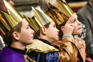 Drei Sternsinger sitzen nach der Gabenprozession im Petersdom und verfolgen den Gottesdienst mit dem Papst. Die Sternsinger haben die Gewänder und Kronen an. Einer der Sternsinger hat den Arm leicht erhoben und deutet mit dem Zeigefinger auf etwas. Einer der anderen Sternsinger folgt mit seinem Blick. 