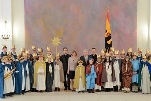 Bundespräsident zusammen mit über 30 Sternsingern. Der Bundespräsident steht zusammen mit seiner Frau und Prälat Krämer in der Mitte. Die Sternsinger stehen recht und links von ihnen. Ein Sternsinger auf linken Seite trägt ein Saxophon um den Hals. Alle Sternsinger haben bunte Umhänge und Gewänder, sowie Goldenen Turbane und Kronen an. 