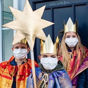 Drei Sternsinger mit Mund-Nasen-Schutz vor einer blauen Haustür.