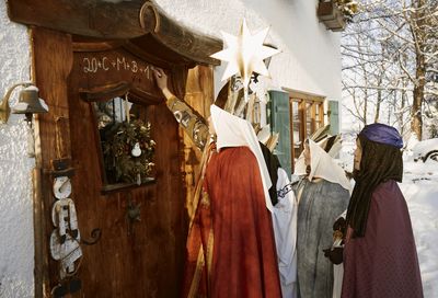 Vier Sternsinger stehen vor einer Hautür. Sie haben die traditionellen Gewänder der Heiligen Drei Könige an. Einer der Sternsinger hat Kreide in der Hand und schreit den traditionellen Segen der Sternsinger an eine Haustür. 