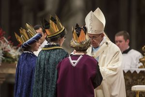 Papst Franziskus blickt die drei Sternsinger an die direkt vor ihm stehen. Die Sternsinger haben Gewänder und Kronen an. Die Sternsinger nehmen an der Messe im Petersdom teil. Sie tragen die traditionelle Kleidung der Heiligen Drei Könige uns glänzende goldene Kronen. 