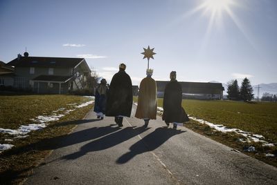 Drei Sternsinger tragen den Stern voran und befinden sich auf dem Weg zu einem Haus. Die Sonne scheint.