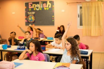 Viele Kinder sitzen in einem Klassenzimmer und lernen. Einige der Kinder zeigen auf um etwas zu sagen. Mädchen und Jungen haben gemeinsam Unterricht. 