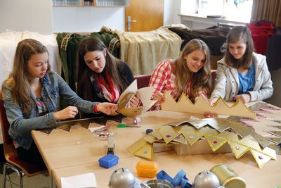 Die Sternsinger Alexandra, Jana, Alina und Carolin sitzen an einem großen Tisch uns basteln Kronen. Sie bereiten sich auf die Sternsingeraktion vor und verzieren die Kronen mit vielen Detailwie glitzerndem Staub und funkelnden Steinen. 