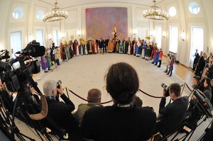 Der Blick von Seiten der Presse. Die Reporter haben Foto- und Fernsehkameras aufgebaut und verfolgen das Geschehen aufmerksam. Bundespräsident Joachim Gauck und seine Frau Daniela stehen zusammen mit dem Prälaten  Dr. Klaus Krämer in der Mitte des Raumes. Auf der linken Seite stehen Sternsinger mit Instrumenten. Diese Sternsinger spielen ein Lied vor. Die Sternsinger auf der rechten Seite singen dazu ein Lied. Der Raum indem alle sind ist groß und hell. Es hängen große Kronleuchter von der Decke herab.