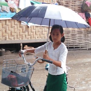 Shwe-San hält einen Regenschirm über sich und sitzt auf einem zu großen Fahrrad.