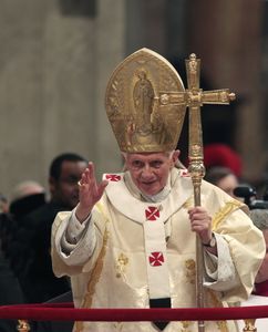 Papst Franziskus segnet die beim Gottesdienst anwesenden Menschen. Er steht aufrecht und hat ein großes goldenen Kruzifix in der linken Hand. Die rechte Hand hat er zum Segen erhoben. Papst Benedikt trägt sein volles Ornat.