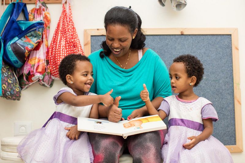 UNITAF Kindergarten und Tagesstaette Auf dem Bild ist ein Zwillingspaar zusammen mit einer Betreuerin zu sehen. Sie sitzen auf dem Boden und Lesen ein Bilderbuch. Alle Lächeln halten die Hand vor den Körper und strecken den den Daumen in die Luft.