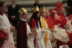 Drei Sternsinger nehmen am Neujahrsgottesdienst im Petersdom teil. Jeder der Drei Sternsinger trägt kleine Gaben und Reliquien indem Händen. Die Sternsinger schreiten grade die Reihen der Kardinäle ab. 