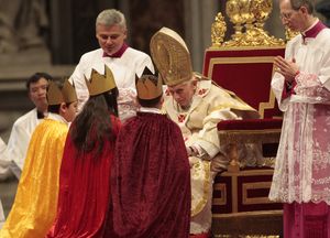 Drei Sternsinger stehen vor Papst Benedikt. Sie haben die traditionellen Gewänder an. Die Sternsinger knien vor dem Papst der in seinem Thron sitzt. Papst Benedikt hat sich in seinem Thron nach vorne gebeugt um besser mit den Sternsingern reden zu können. 