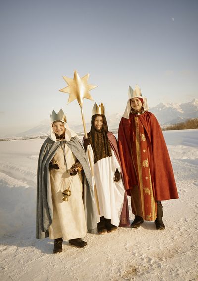 Drei Sternsinger stehen mit den langen Gewändern der Heiligen Drei Könige im Schnee. Am Horizont sind schneebedeckte Berge. Die Sternsinger tragen Kronen, haben Weihrauch dabei und tragen den Stern. 