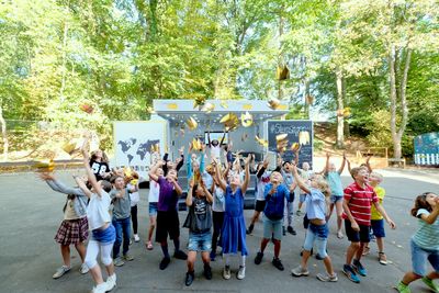 Viele Kinder springen hoch und werfen goldene Papierkronen in die Luft. Im Hintergrund ist ein ein weißer Transporter zu sehen, der ausgeklappt ist.