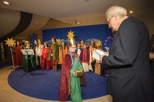 Der deutsche EU-Vizepräsident Rainer Wieland spendet etwas an die Sternsinger. Vor ihm steht eine Sternsingerin aus Belgien mit einer Sternsinger Spendenbox. Rainer Wieland wirft seine Spende in diese Spendenbox. Im Hintergrund stehen die restlichen neunzehn Sternsinger und gucken den Beiden zu.