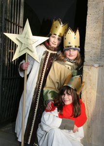 Drei Sternsinger stehen in einem Hauseingang und blicken in die Kamera. Sie tragen goldene Kronen und lange Umhänge in weiß, braun und rot. Die drei haben auch den Stern der Sternsinger an einem langen Stock dabei.