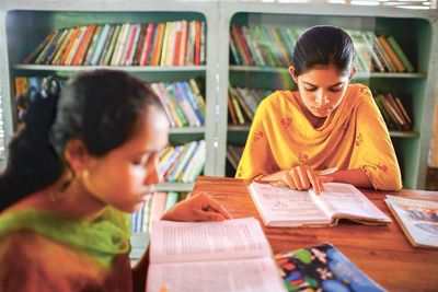 Zwei Mädchen in traditioneller indischer Kleidung sitzen an einem Tisch und lesen Bücher. Im Hintergrund sind Regale die voll mit Büchern sind. Die Mädchen sind konzentriert und lernen für die Schule.