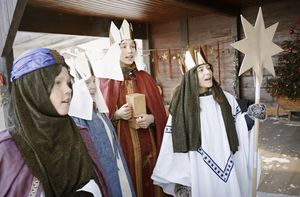 Drei Sternsinger stehen vor einer Haustür und singen. Sie tragen die Gewänder der Heiligen Drei Könige, die Kronen, den Stern von Betlehem und Singen.
