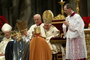 Drei Sternsinger knien vor dem Papst. Sie haben die traditionellen Gewänder an und tragen die goldenen Kronen auf dem Kopf. Der Papst segnet die Sternsinger. Um das besser tun zu können hat er sich in seinem Stuhl weiter nach vorne gebeugt.