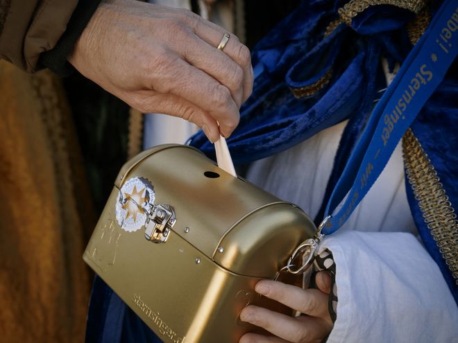 Ein Sternsinger hält die goldene Spendenbox in den Händen. Eine andere Person wirft gerade eine Spende in diese Box. Die Box ist mit einem Sternsingeraufkleber versiegelt.