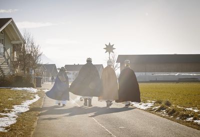 Vier Sternsinger auf dem Weg zu einem Gehöft. Sie gehen in strahlendem Sonnenschein einen Schotterweg entlang. Alle gehen nebeneinander und den Stern haben sie in der Mitte.