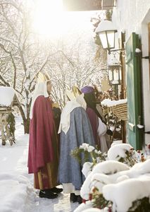 Vier Sternsinger stehen in voller Montur in einem Hauseingang und singen. Es liegt hoher Schnee und die Sonne scheint.