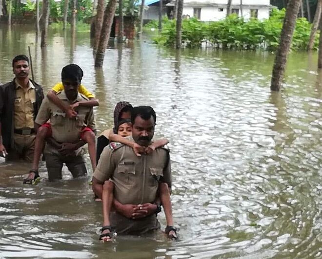 Bild aus den Überschwemmungsgebieten in Indien. Helfer tragen Kinder und Jugendliche aus den Gefahrengebieten.  Das Wasser steht den ausgewachsenen Männern in Uniform bis zur Taille. Im Hintergrund sind Häuser zu sehen, die überschwemmt worden sind. 