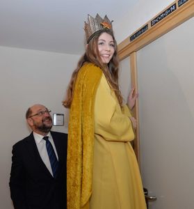 Lena aus Österreich schreibt den traditionellen Segen der Sternsinger über die Tür des EU- Parlamentspräsidenten Martin Schulz. Sie trägt einen leuchtend gelben Umhang und hat eine Krone auf dem Kopf. Den Segen hat sie mit Kreide geschrieben. Martin Schulz blickt glücklich auf den Segen auf seiner Tür. Der Segen lautet CMB. Das heißt Christus segne dieses Haus.