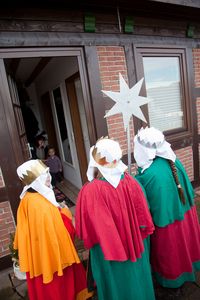 Drei Sternsinger stehen an der Tür einer Hütte und geben ein Sternsingerlied zum Besten. Die Sternsinger haben die traditionellen Gewänder der Heiligen Drei Könige an.