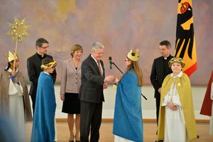 Bundespräsident Joachim Gauck übergibt einer Sternsingerin eine Spende. Die Sternsingern hält die Spendenbox vor sich und Joachim Gauck wirft seine Spende in die Spendenbox. Im Hintergrund stehen seine Frau Daniela Schadt und Prälat Dr. Klaus Krämer