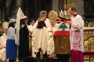 Vier Sternsinger sind vorne beim Papst und unterhalten sich mit Ihm. Die Sternsinger haben die Gewänder und Kronen der Heiligen Drei Könige an. Einer der Sternsinger hat die Nationalflagge des Libanon auf den Rücken. Die Flagge ist rot und weiß und hat und er Mitte eine grüne Tanne. 