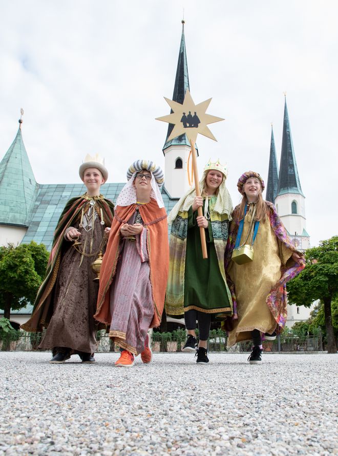 Die vier Sternsinger sind gekleidet in die Gewänder der Heiligen Drei Könige und tragen den Stern von Betlehem vor sich her. Die Sternsinger befinden sich vor der Gnadenkapelle in Altötting. Die Kapelle ist im Rücken der Sternsinger und die Vier gehen auf die Kamera zu.