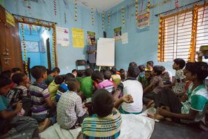 4_Tag gegen ausbeuterische Kinderarbeit Eine Einblick in den informellen Unterricht der durch ARKTF ermöglicht wird. Hier werden Kinder für eine Stunde am Tag im Rechnen, Lesen und Schreiben unterrichtet. In dem Bild sind über 31 Kinder zu sehen die einer Lehrerin zuhören. Die Kinder haben keine Schreibtische oder Stühle. Sie sitzen auf dem Boden und hören zu. Sie haben keine Hefte oder Stifte. Die Länder des Klassenraums sind freundlich Blau angestrichen. Das Klassenzimmer ist voll, wirkt dennoch luftig und freundlich.