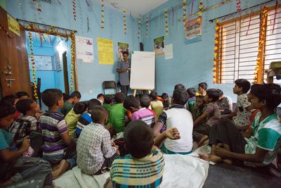 Eine Einblick in den informellen Unterricht der durch ARKTF ermöglicht wird. Hier werden Kinder für eine Stunde am Tag im Rechnen, Lesen und Schreiben unterrichtet. In dem Bild sind über 31 Kinder zu sehen die einer Lehrerin zuhören. Die Kinder haben keine  Schreibtische oder Stühle. Sie sitzen auf dem Boden und hören zu. Sie haben keine Hefte oder Stifte. Die Länder des Klassenraums sind freundlich Blau angestrichen. Das Klassenzimmer ist voll, wirkt dennoch luftig und freundlich. 