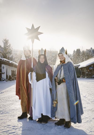 Diese dreiköpfige Gruppe der Sternsinger ist gekleidet in lange Umhänge und glitzernde Kronen. Sie tragen lila farbene, blaue und rote Umhänge. Die Gruppe steht im Schnee und die Sonne steht schon tief am Himmel. Der Stern der Sternsinger strahlt weil die Lichtstrahlen den Stern von hinten erleuchten.