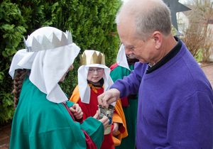 Drei Sternsinger berichten zufrieden wie ein Mann eine Spende in die Spendendose wirft. Die Sternsinger haben die Gewänder und Kronen an.