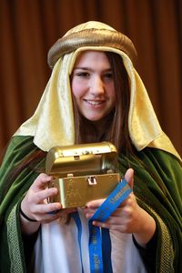 Eine Sternsingerin in einem grünen Umhang mit goldenen Verzierungen an den Ärmeln hält mit beiden Händen eine Sternsinger Spendenbox. Die Spendenbox ist golden und hat auch ein Schloss um die Box sicher zu verschließen. 