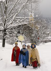 Vier Sternsinger auf ihrem Weg durch das verschneite Dorf. Die Äste an den Bäumen sind dick von Schnee bedeckt genau so wie die umliegenden Häuser. Die Sternsinger  tragen rote, blaue und beige Umhänge und ahn auch die Kronen auf dem Kopf. Zudem tragen sie den Stern der Sternsinger an einem langen Stock mit sich.