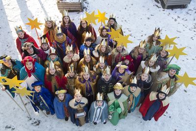 Eine Gruppe von 35 Sternsingern stehen im Schnee. Sie haben die bunten Gewänder der Heiligen Drei Könige an. Sie tragen goldene Kronen und halten Sterne in die Luft.