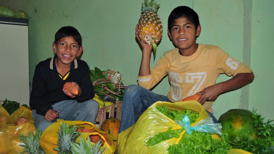 Jonathan hilft beim waschen, einkaufen, schneiden und kochen von Gemüse. Hier hält er eine Knolle in die Luft, sein Freund hält eine Ananas.