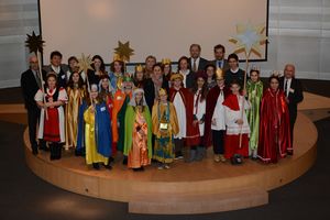 Die Sternsinger aus den verschiedenen europäischen Ländern stehen zusammen auf einem hölzernen Podest. Die Betreuer stehen auch dort. Die Sternsinger tragen bunte Umhängend goldenen Kronen. Im Hintergrund ist Vorstandsmitglied des Kindermissionswerks Dr. Franz Marcus zu sehen. Die Sternsinger halten insgesamt vier goldene Sterne an langen stecken aus Holz in die Höhe.