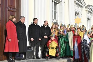 Mehr als zwanzig Sternsingern stehen auf der Treppe von Schloss Bellevue und werden von Bundespräsident Joachim gack und seiner Lebensgefährtin begrüßt. Die Sternsinger sind festlich mit vielen Farben gekleidet und tragen glänzende Kronen auf dem Kopf. einer der Sternsinger sitzt im Rollstuhl. Prälat Krämer steht neben dem Bundespräsidenten.